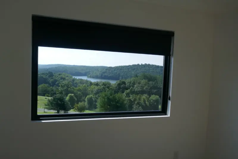 Black roller shade half lowered on a picture window with a lake and treeline view