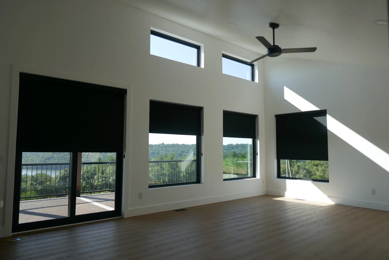 Dark blackout roller shades on five windows of a modern living room with a ceiling fan
