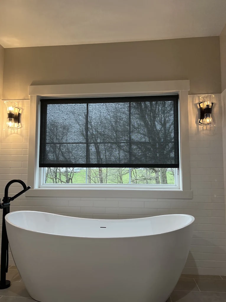 Dark solar screen shade over a bathroom window above a freestanding tub, trees visible through the weave