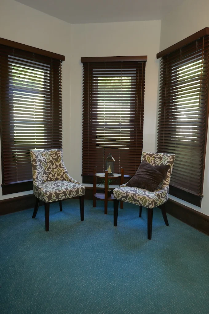 Dark wood blinds on three bay windows behind two patterned armchairs on teal carpet