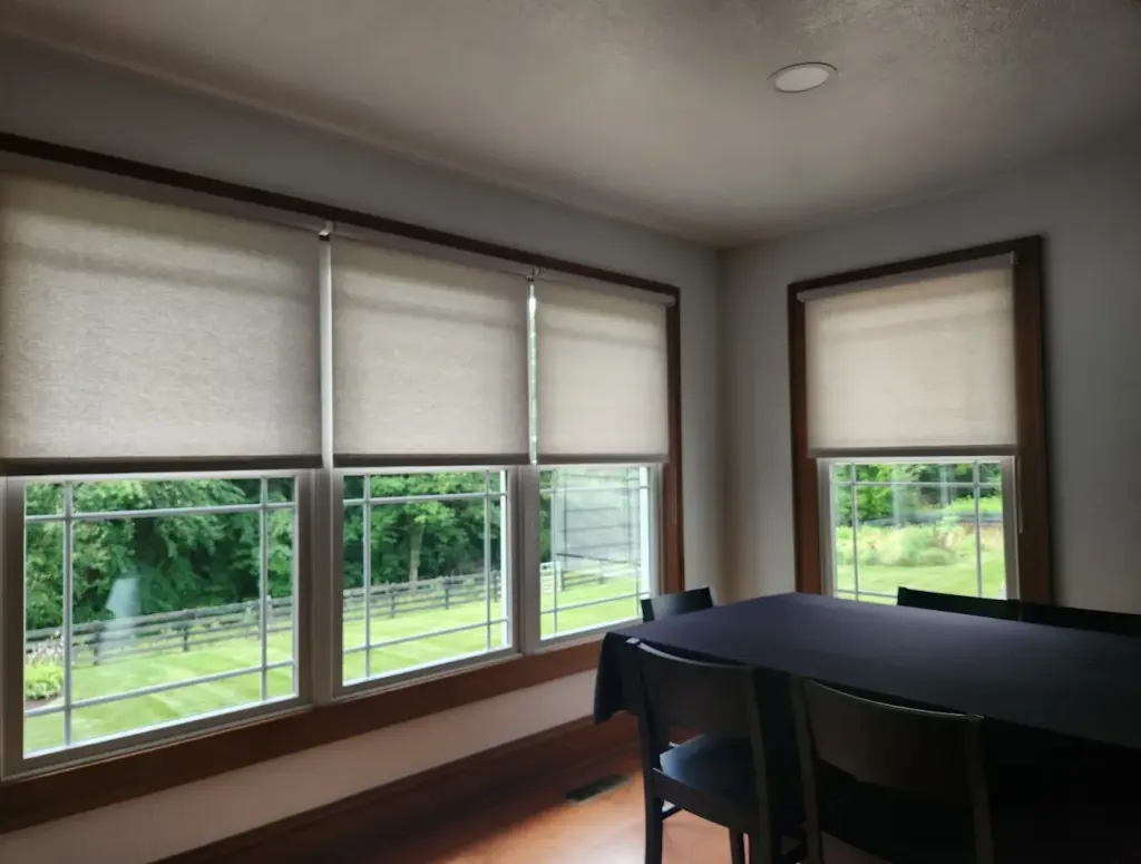 Grey light-filtering roller shades on two wood-trimmed dining room windows with a view of fields