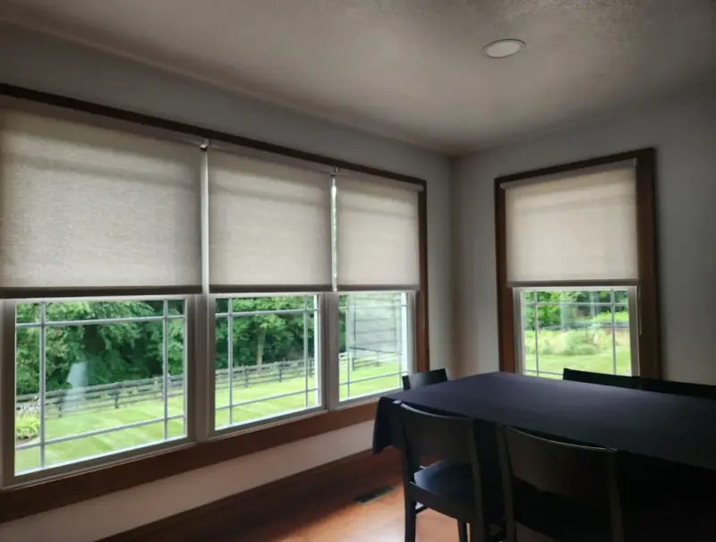 Grey light-filtering roller shades on two wood-trimmed dining room windows with a view of fields