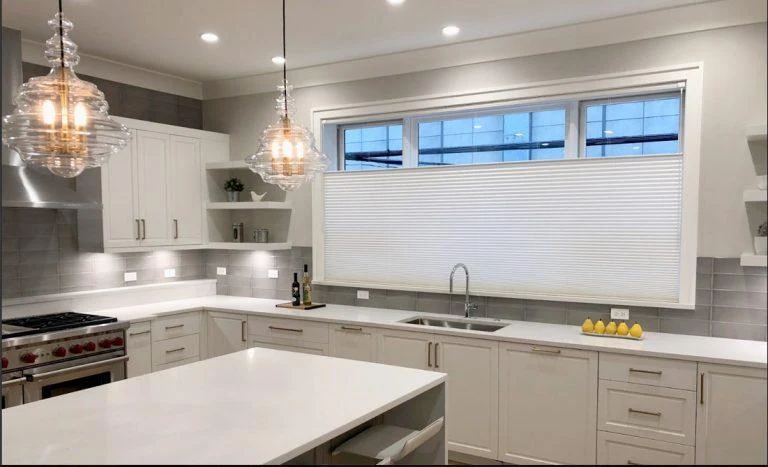 White cellular shade over a kitchen sink window with pendant lights and a stone island