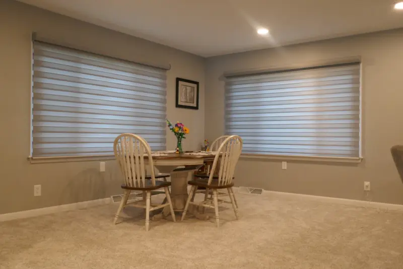 Zebra shades on two windows of a carpeted dining room with a round table